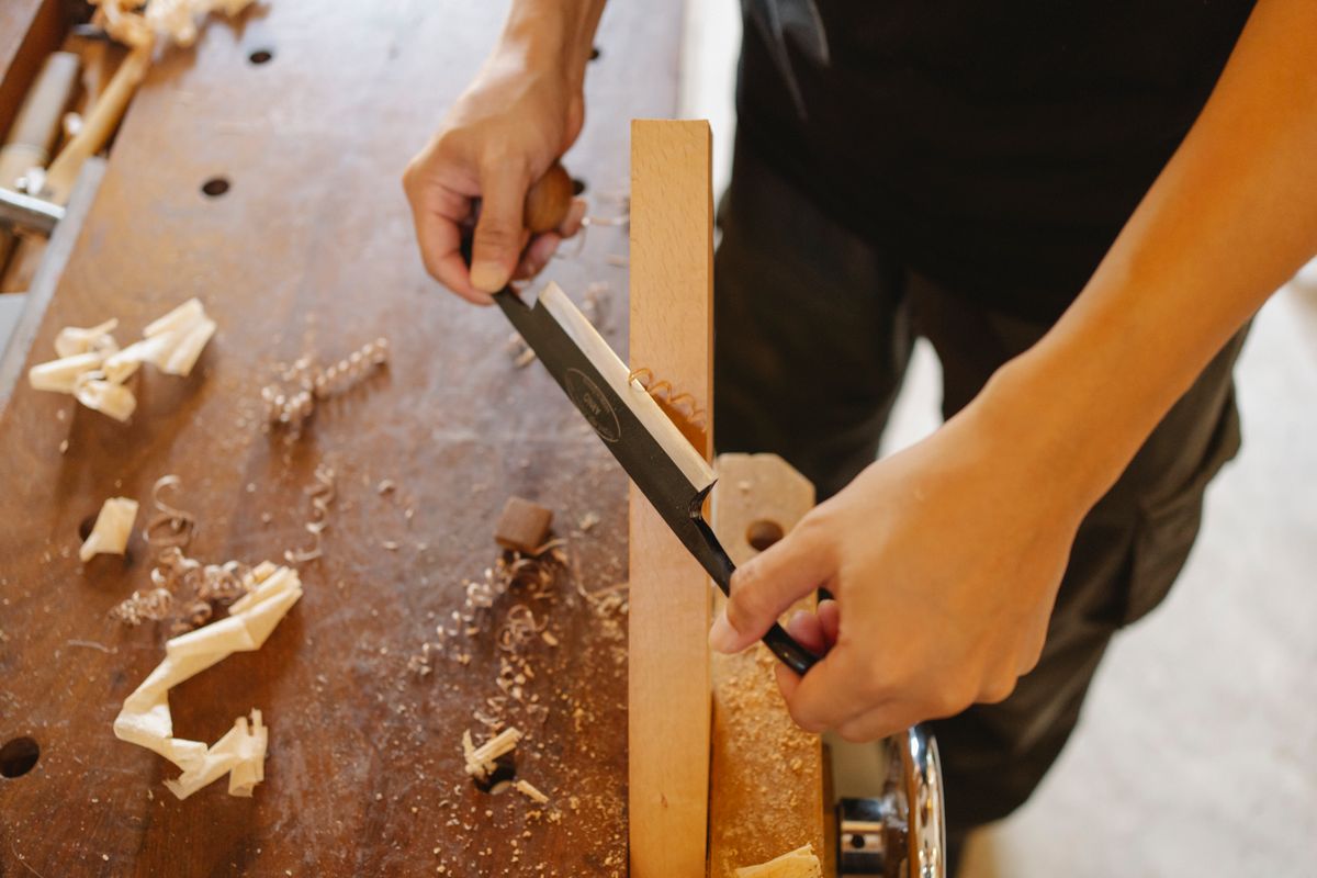 Two hands paring a tenon shoulder with a bench chisel, shavings across the bench top