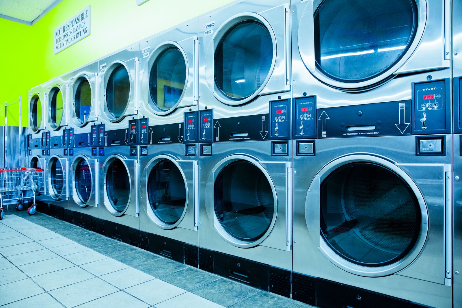 A bright laundromat: rows of front-loaders along a green wall, folding tables beyond