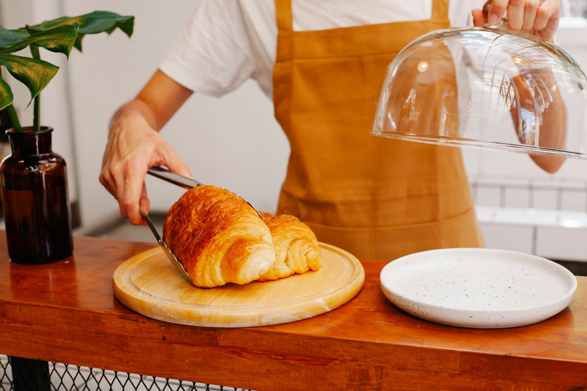 A staff member lifting the glass cloche to lift a croissant onto a plate with tongs