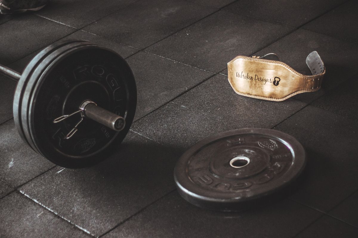 A leather lifting belt on the floor beside a loaded bar and a spare plate
