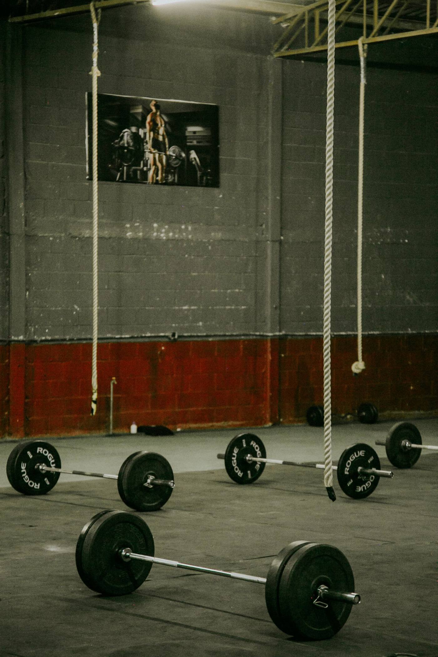 An industrial gym floor: loaded barbells, plates racked along the wall, ropes overhead