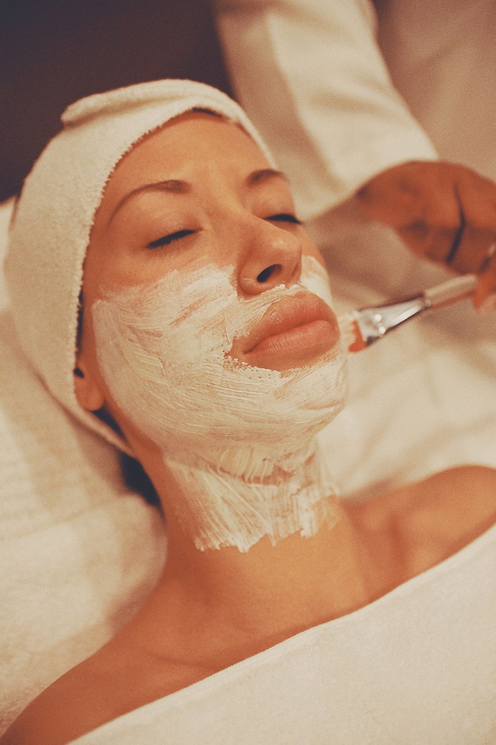 A treatment mask being applied with a brush while the client rests under a warm towel