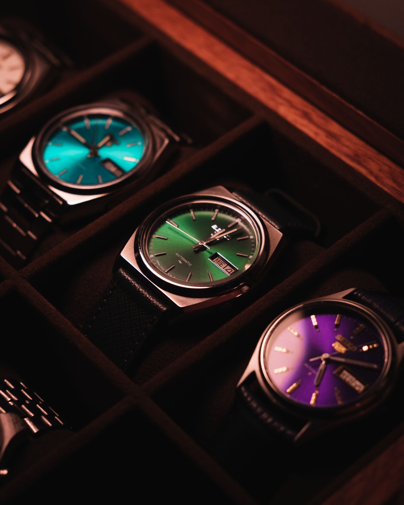 Four watches lying in the felt trays of an open watch case, dials in teal, green and purple