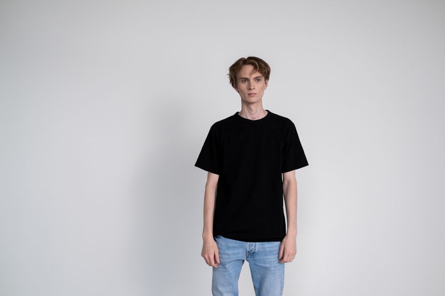 A figure in a plain black heavyweight tee and jeans against a pale studio wall