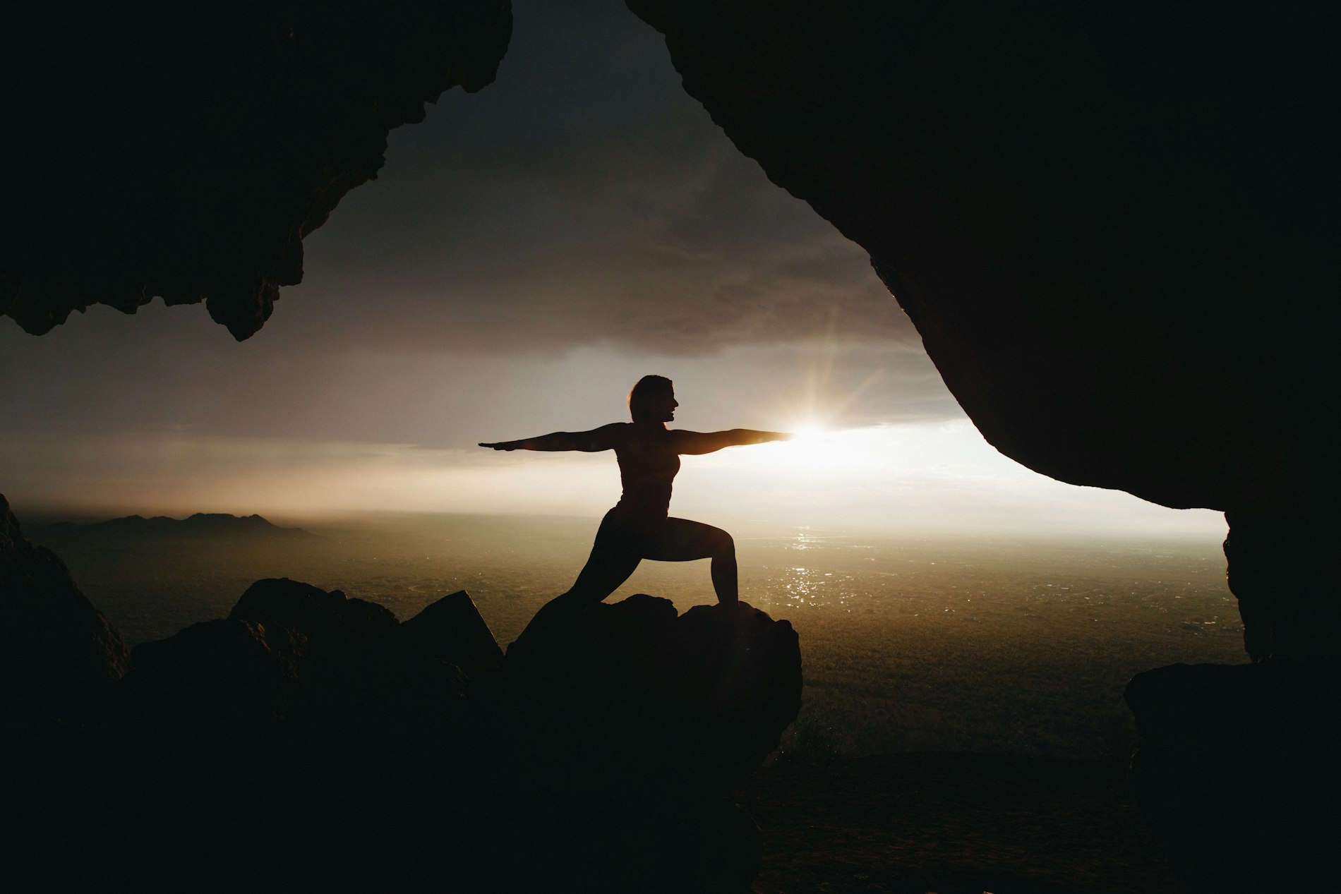 A person holding warrior pose on a rock ledge at sunset, silhouetted against the light over the valley