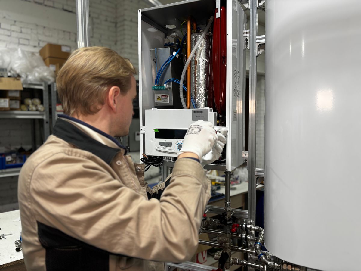 A technician working inside the open front of a wall-mounted boiler beside a hot water cylinder