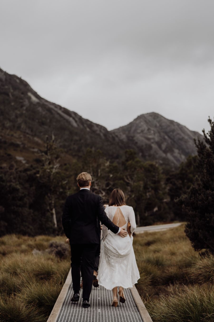 A couple in wedding clothes walking a narrow boardwalk toward grey mountains under low cloud