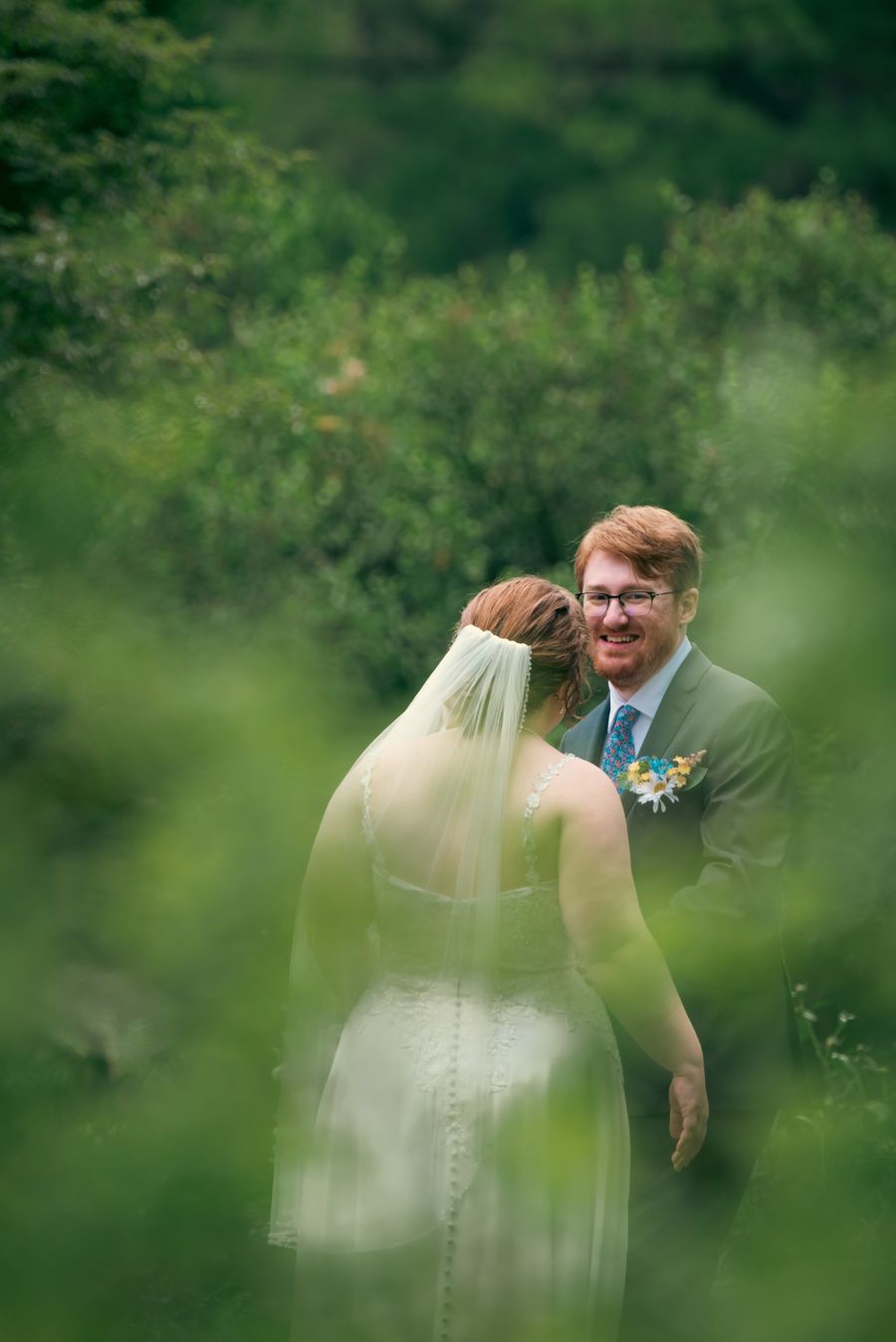 A groom seeing his bride for the first time, both half-hidden by summer leaves