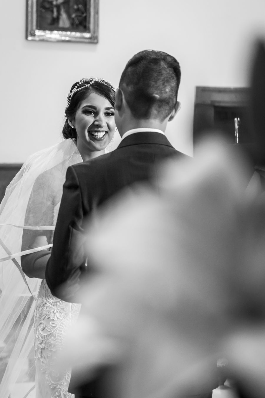 Black and white photograph of a bride laughing during the ring exchange, shot past a foreground of flowers
