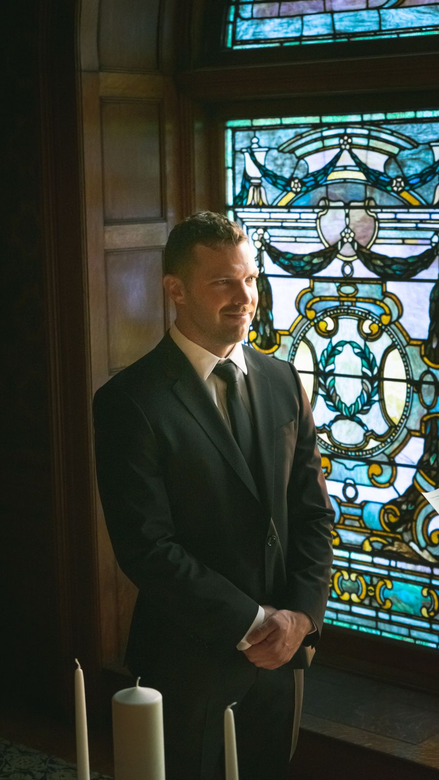 A groom waiting alone beside a tall stained-glass window in a dim chapel